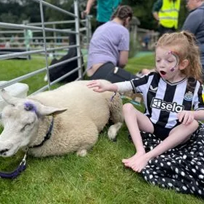 Little girl sitting outside on a blanket with a sheep to her side.